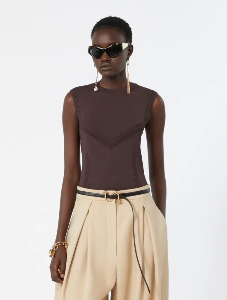 Model wearing a sleeveless brown top with chevron stitch details and beige wide-leg trousers, with black sunglasses and gold jewelry.