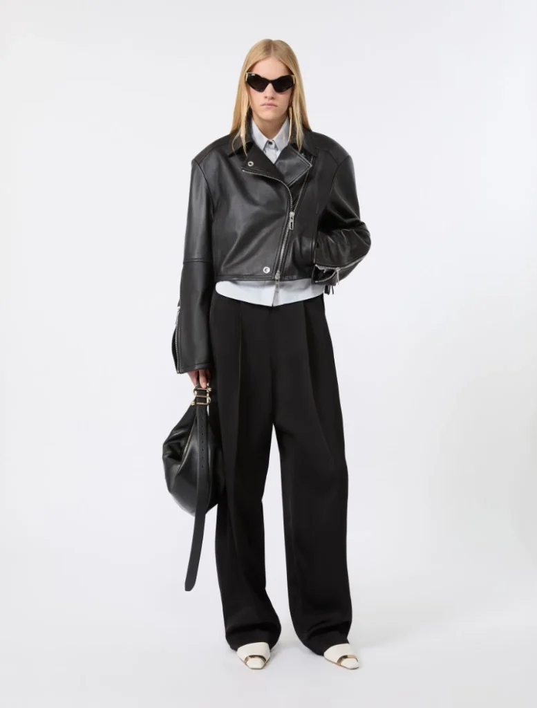 Model in a cropped black leather jacket, white shirt, and wide-leg black trousers with dark sunglasses and a black handbag.