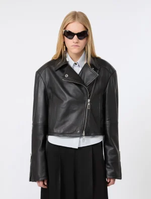 Model wearing a black biker leather jacket over a light shirt and black skirt with dark sunglasses, posed against a plain backdrop.