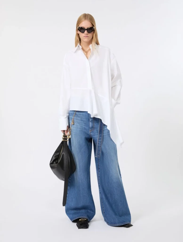 Woman in oversized white shirt with wide-leg blue jeans, black handbag, and dark sunglasses, posing against a white studio backdrop.
