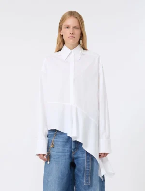 Model wearing an oversized white shirt with an asymmetric hem over blue jeans, standing in a neutral studio setting. The shirt features a structured collar and long sleeves with wide cuffs; a gold-toned chain accessory hangs from the jeans.