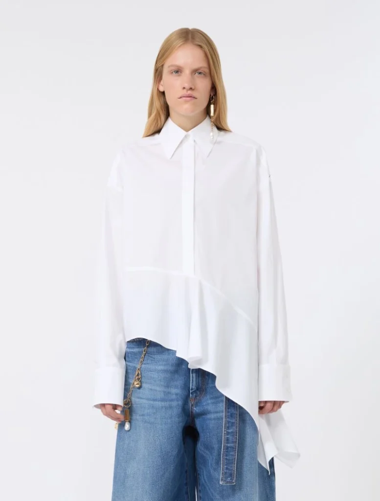 Model wearing an oversized white shirt with an asymmetric hem over blue jeans, standing in a neutral studio setting. The shirt features a structured collar and long sleeves with wide cuffs; a gold-toned chain accessory hangs from the jeans.