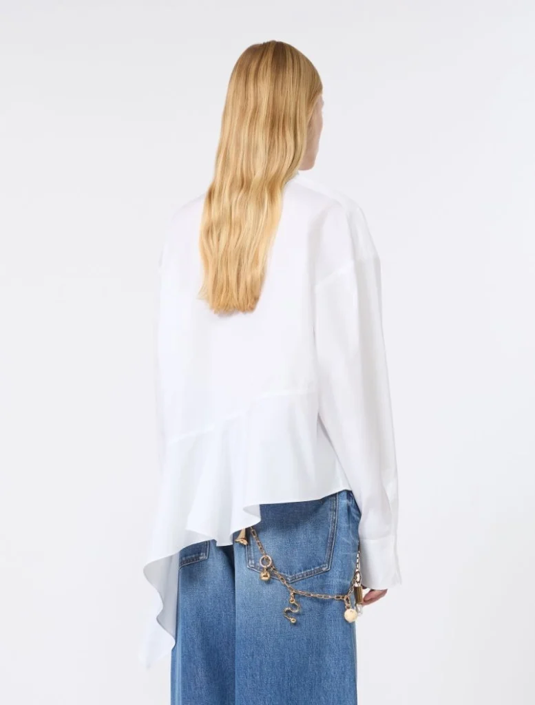 Back view of a person with long blonde hair wearing a white asymmetrical blouse and blue denim jeans with a gold chain accessory on the pocket area?