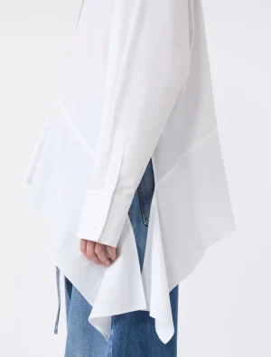 Side view of a loose white shirt with an asymmetrical, draped hem over blue jeans.