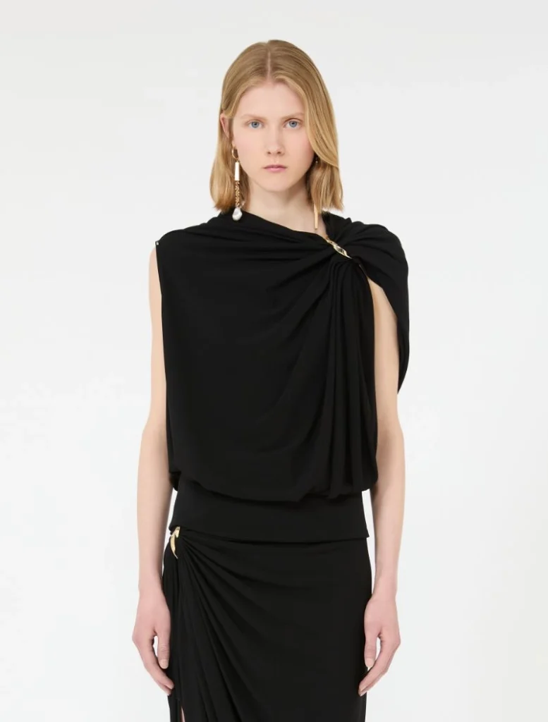 Model wearing a black draped one-shoulder top and matching black skirt with gold jewelry.