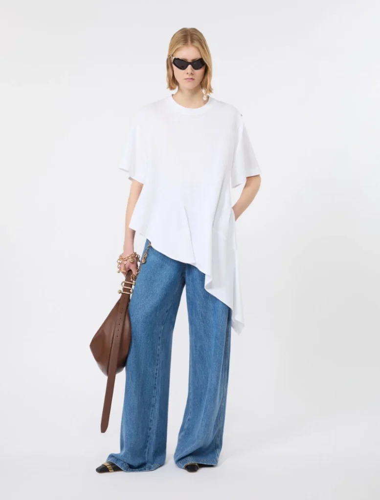 Model in oversized white asymmetrical top with wide-leg blue jeans, black sunglasses, brown shoulder bag, and flat sandals.