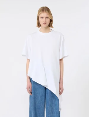 Woman wearing an oversized white asymmetric top over wide-leg blue jeans, standing against a plain light background.