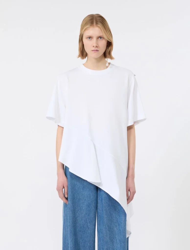 Woman wearing an oversized white asymmetric top over wide-leg blue jeans, standing against a plain light background.