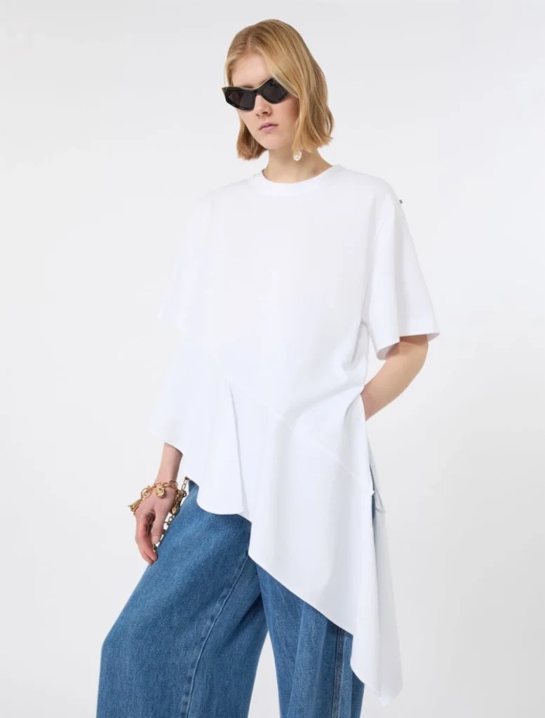 Model wearing an oversized white tunic with an asymmetric, draped hem over wide-leg blue jeans, accessorized with black sunglasses.