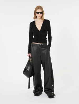 Model wearing a black ruched V-neck top with wide-leg leather pants, carrying a large black handbag and wearing sunglasses.