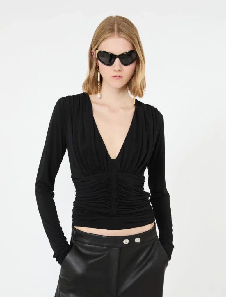 Model wearing a black ruched V-neck top with black leather pants, accessorized with oversized sunglasses and dangling earrings.