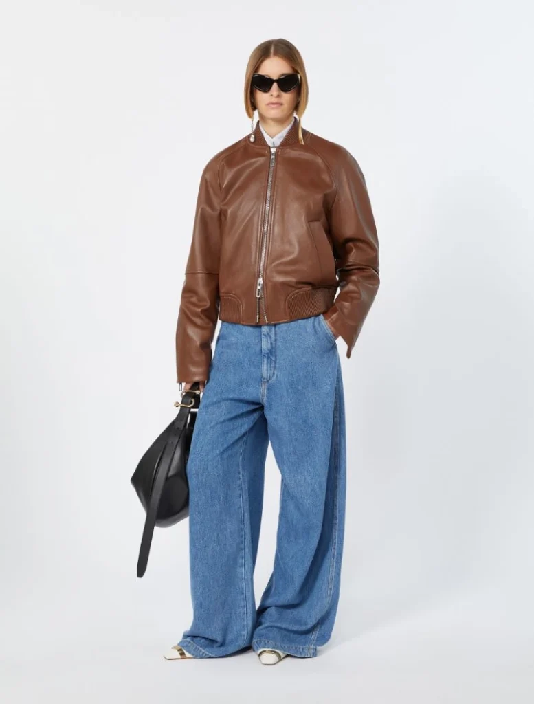 Model wearing a brown leather bomber jacket with wide-leg blue jeans, white shirt, black sunglasses, and a black handbag.