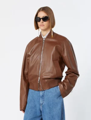 Woman wearing a brown leather bomber jacket, blue jeans, and oversized black sunglasses against a white background.