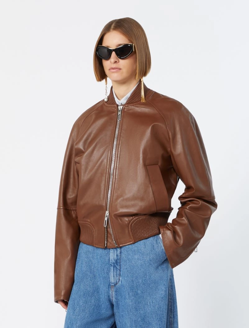 IMG_3080 - New Trend Woman wearing a brown leather bomber jacket, blue jeans, and oversized black sunglasses against a white background.