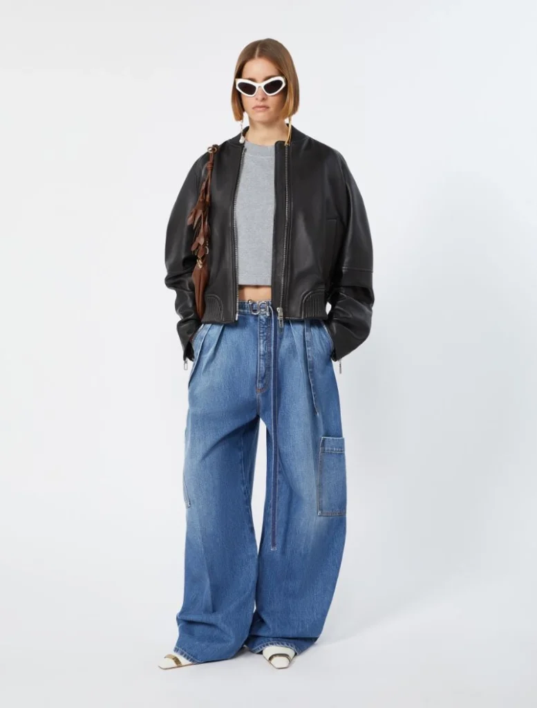 Full-body shot of a model wearing a black bomber jacket, gray crop top, and oversized blue jeans with a brown handbag and white sunglasses.",