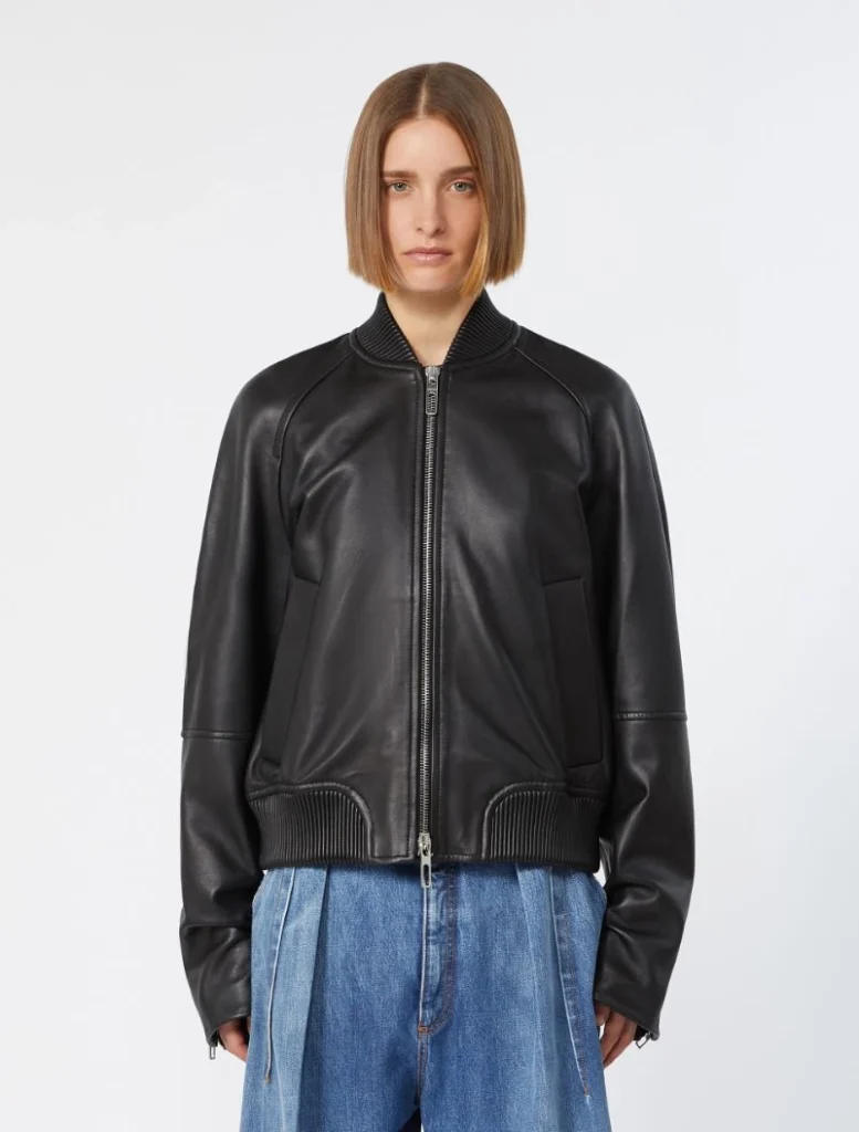 Woman wearing a black leather bomber jacket with a front zipper and ribbed cuffs, paired with blue jeans in a studio setting.