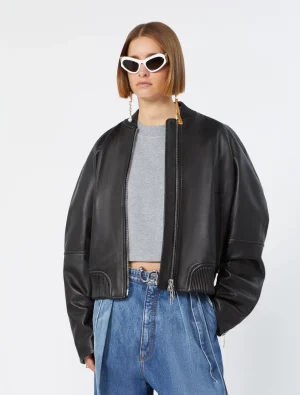 Woman wearing a black leather bomber jacket over a gray crop top with blue jeans and white cat-eye sunglasses.