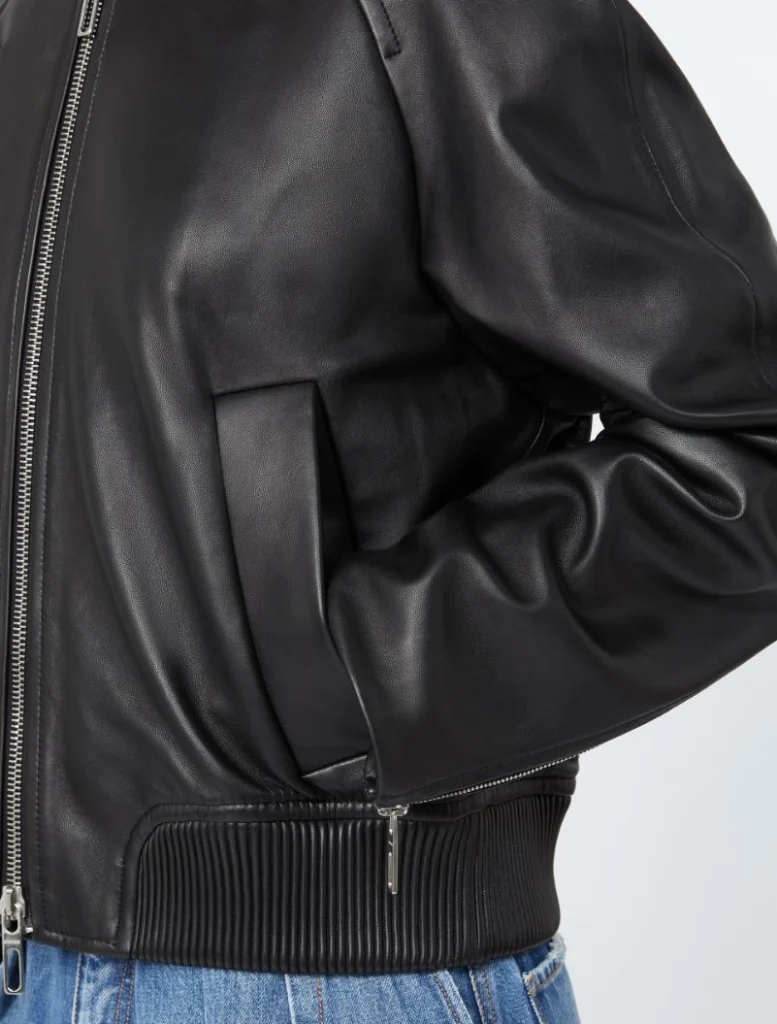Close-up of a black leather bomber jacket with a zip front, pocket, and ribbed hem, worn over blue jeans