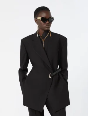 Model wearing an oversized black blazer with a belt, black trousers, statement gold jewelry, and dark sunglasses on a light background.