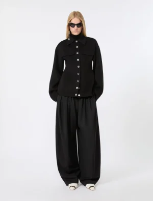 Full-body shot of a woman in an all-black outfit: button-front jacket with large pockets and wide-leg trousers, white sandals, and sunglasses.