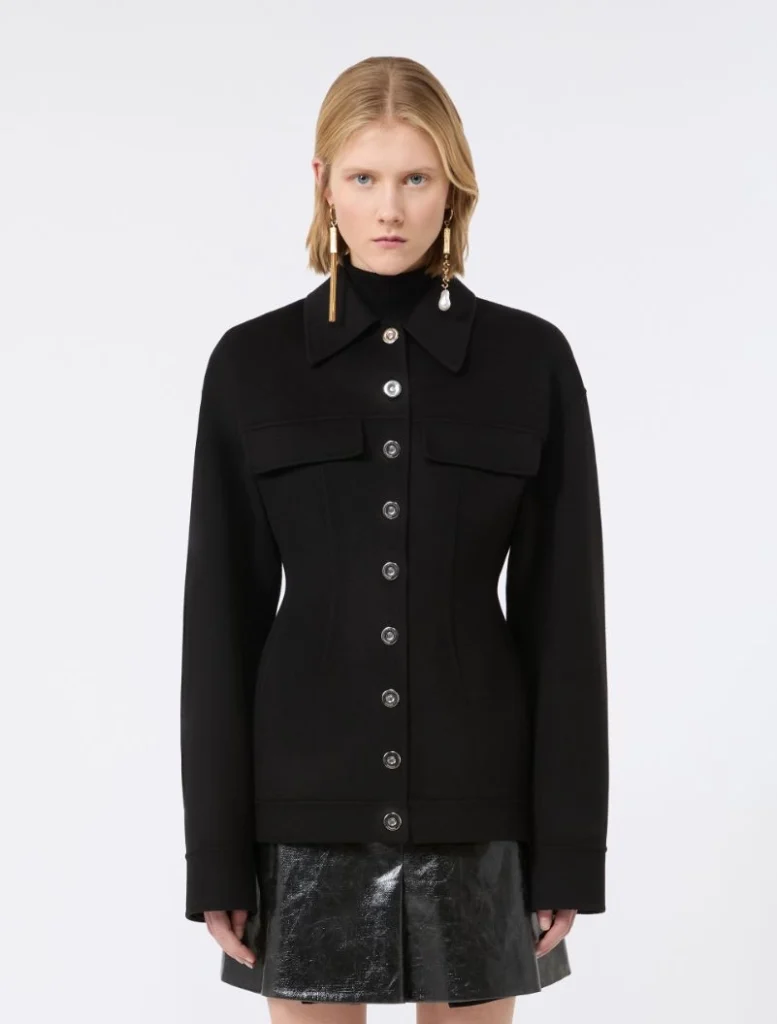 Model wearing a black button-up jacket with wide collar and front flap pockets, paired with a shiny black mini skirt.