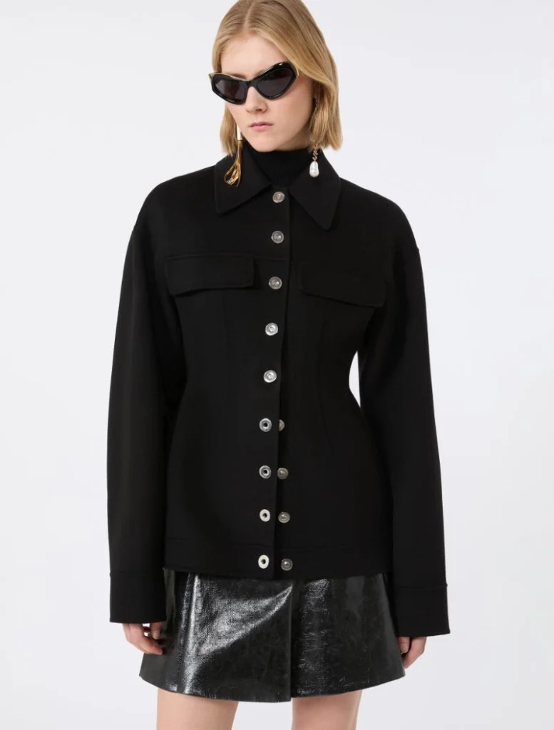 Model wearing a black button-front jacket with large chest pockets, silver buttons, and a shiny black mini skirt, plus sunglasses and earrings.