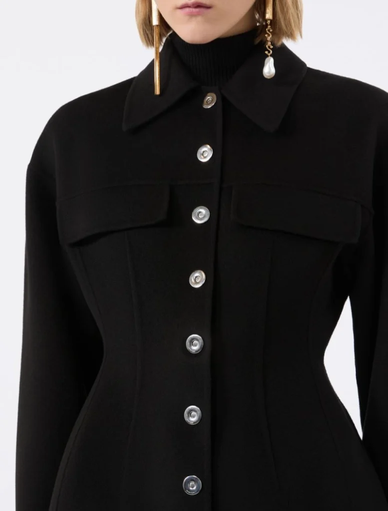 Close-up of a black tailored coat with silver front buttons, worn over a black turtleneck; gold drop earrings visible at the sides.