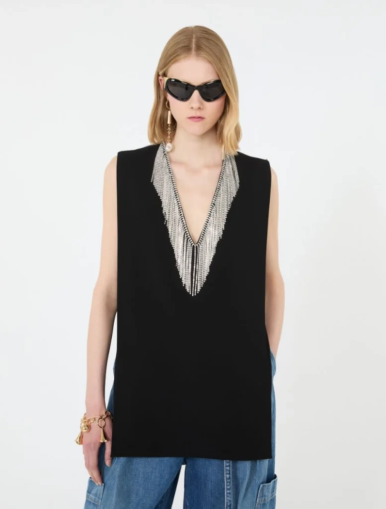 Model wearing a black sleeveless top with a silver beaded V-neck necklace and blue jeans, sunglasses on a white background.