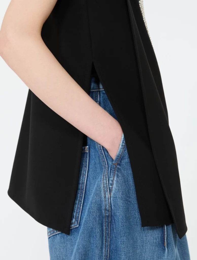 Person wearing a black sleeveless top or vest with blue jeans, left hand in a jeans pocket.