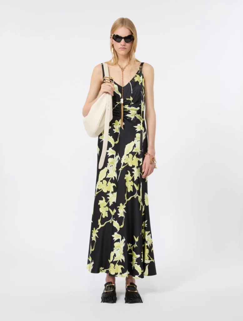 Woman in a black floral maxi dress with pale yellow-green leaves, carrying a cream tote bag and wearing sunglasses.