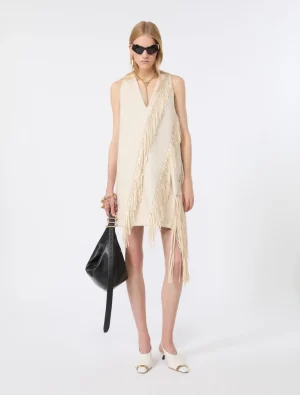 Model wearing a cream one-shoulder fringe dress with a black handbag and white heeled sandals, sunglasses, against a white backdrop.