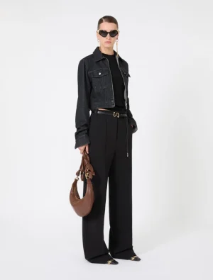 Model wearing a cropped dark denim jacket over a black top, wide-leg black trousers, and black sunglasses, accessorized with a brown crescent-shaped handbag and gold jewelry.