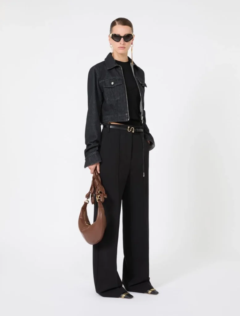 Model wearing a cropped dark denim jacket over a black top, wide-leg black trousers, and black sunglasses, accessorized with a brown crescent-shaped handbag and gold jewelry.
