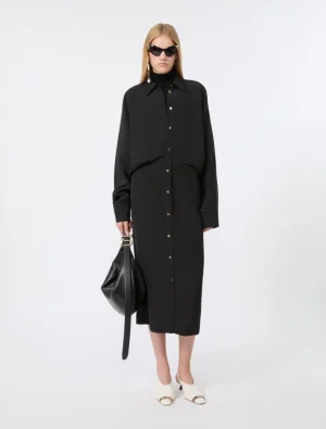 Woman in a long black button-down shirt dress with oversized collar, wearing black sunglasses and carrying a black handbag, white mules.