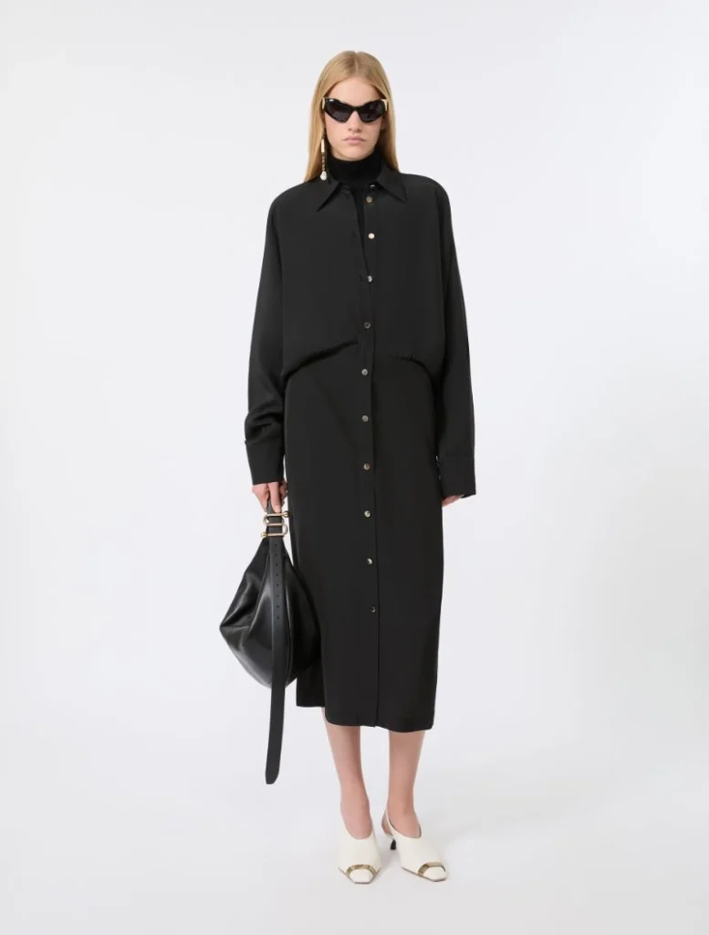 Woman in a long black button-down shirt dress with oversized collar, wearing black sunglasses and carrying a black handbag, white mules.