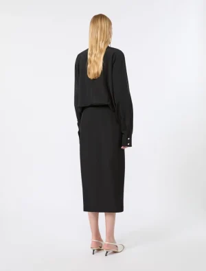 Back view of a woman in an all-black outfit (long-sleeve top and midi dress/skirt) with blonde hair, standing in a white studio setting.