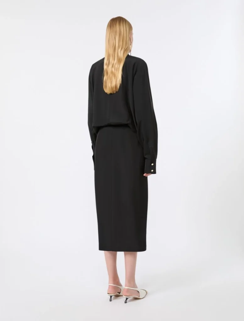 Back view of a woman in an all-black outfit (long-sleeve top and midi dress/skirt) with blonde hair, standing in a white studio setting.