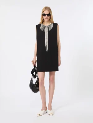 Model wearing a black sleeveless shift dress with a silver-beaded neckline, white heeled mules, oversized sunglasses, and a black handbag.