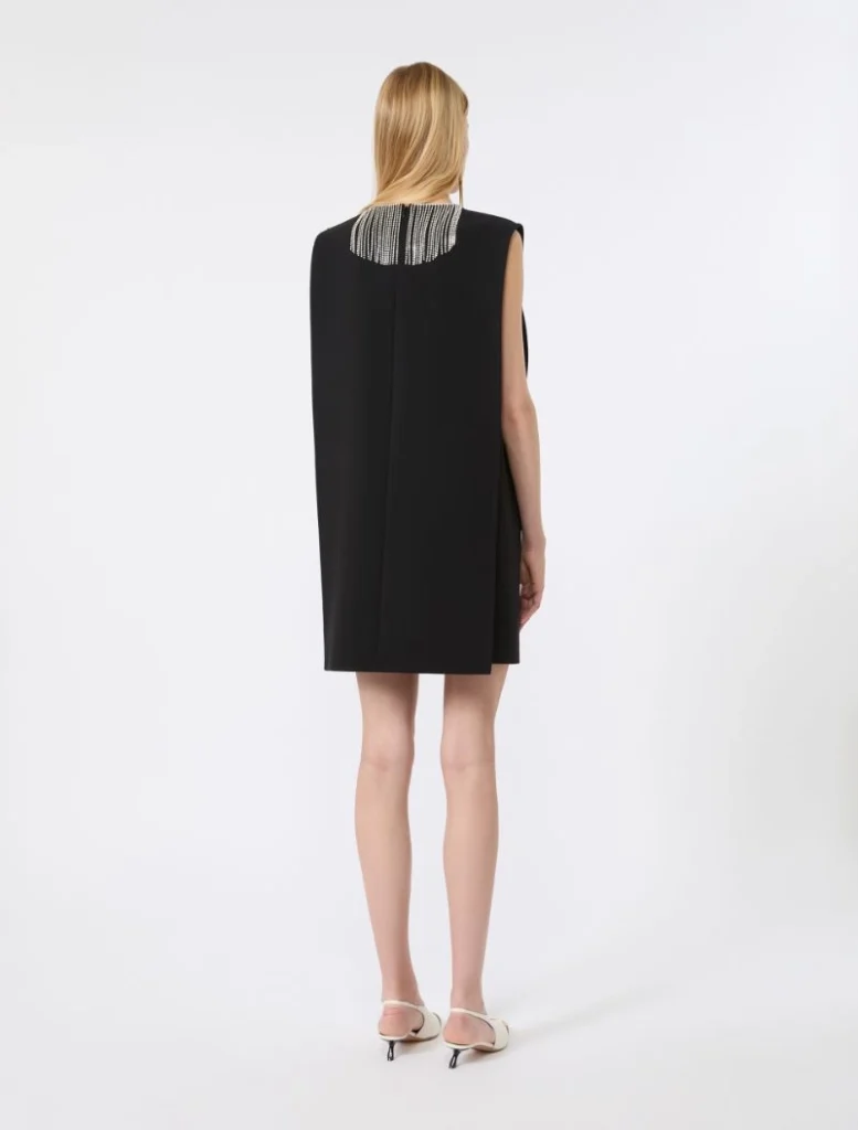 Back view of a model wearing a black sleeveless mini dress with a beaded collar, paired with white strappy heels,