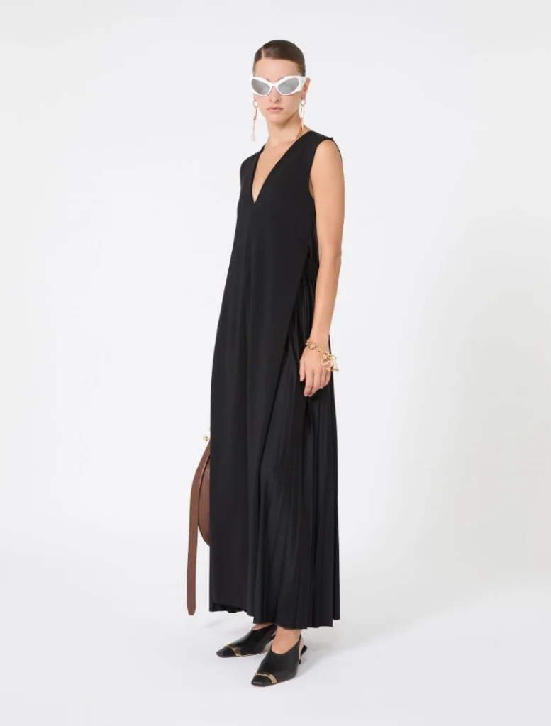 Model wearing a sleeveless black V-neck maxi dress with pleats, white cat-eye sunglasses, gold jewelry, and black gold-toe mules; holding a brown belt/strapbag set on white background.