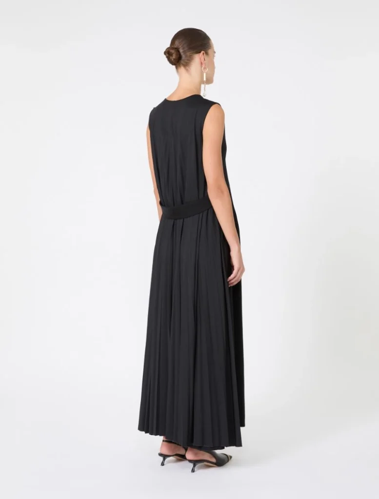 Back view of a woman in a black sleeveless maxi dress with a wide belt, her hair in a bun, wearing dangling earrings and black heeled sandals