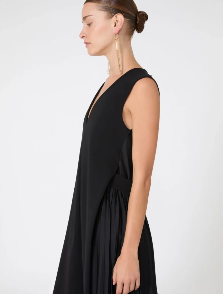 Side profile of a woman wearing a black sleeveless evening dress with a draped overlay and a gold tassel earring.
