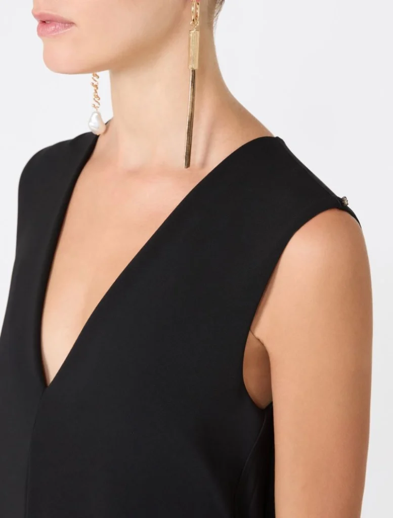 Close-up of a woman wearing a black sleeveless V-neck dress and a long gold bar drop earring with a smaller tassel accent.