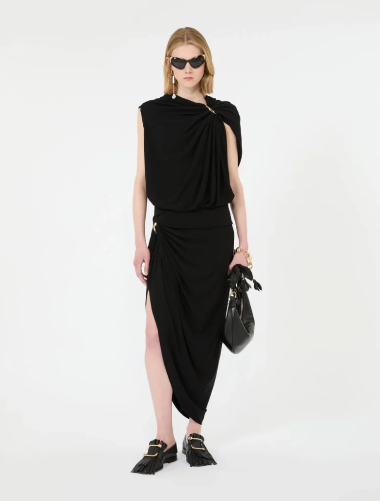 Model wearing a black draped wrap dress with a high side slit, gold jewelry, sunglasses, and black tassel loafers with gold hardware.