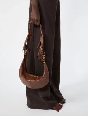 Brown crescent-shaped leather handbag with tassel details held by a person in dark brown pants