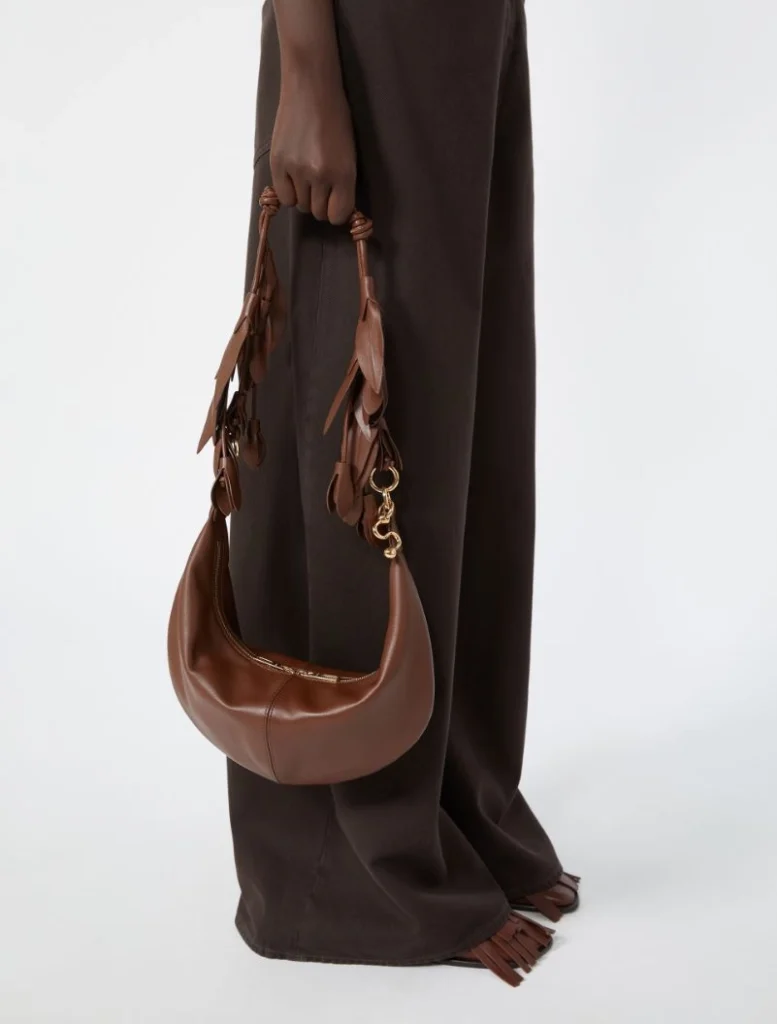 Brown crescent-shaped leather handbag with tassel details held by a person in dark brown pants