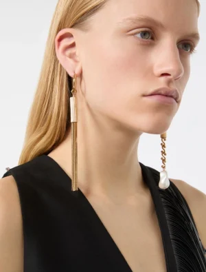 Close-up of a woman wearing a long gold tassel earring on one ear and a chain earring with a pearl on the other, against a black outfit.