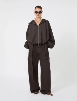 Full-length shot of a model in a dark brown oversized zip-up jacket and wide-leg cargo pants, wearing sunglasses and gold jewelry.