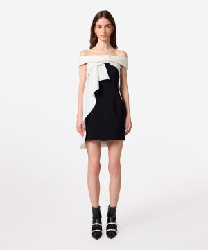 Model wearing a black mini dress with a white off-the-shoulder draped sash and black ankle boots.