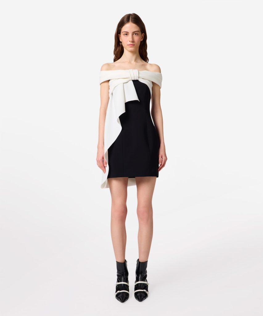 Model wearing a black mini dress with a white off-the-shoulder draped sash and black ankle boots.